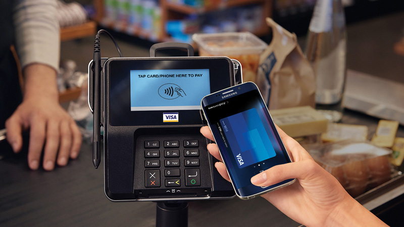 A hand holding a mobile phone showing a Visa card on the screen while in front of a payment terminal.